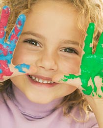 photo of a smiling young girl holding up her messy hands, covered in paint